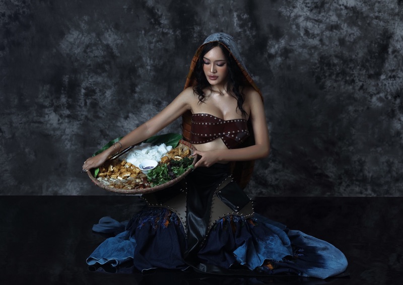 Model in a studded leather bustier and tiered indigo gown holding a woven tray of bun dau mam tom